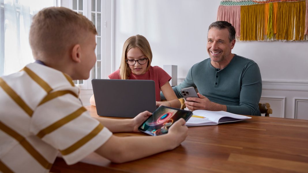 a girl sits at a table on a laptop, a man is next her holding a cellphone and across the table is a young boy holding a tablet