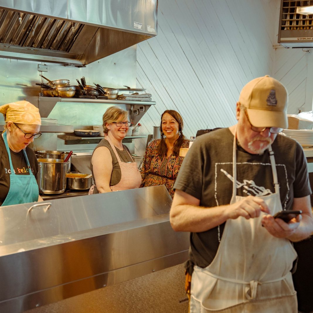 Chris Hyde stands in a kitchen, writing something on a notepad, three woman stand behind him behind a counter