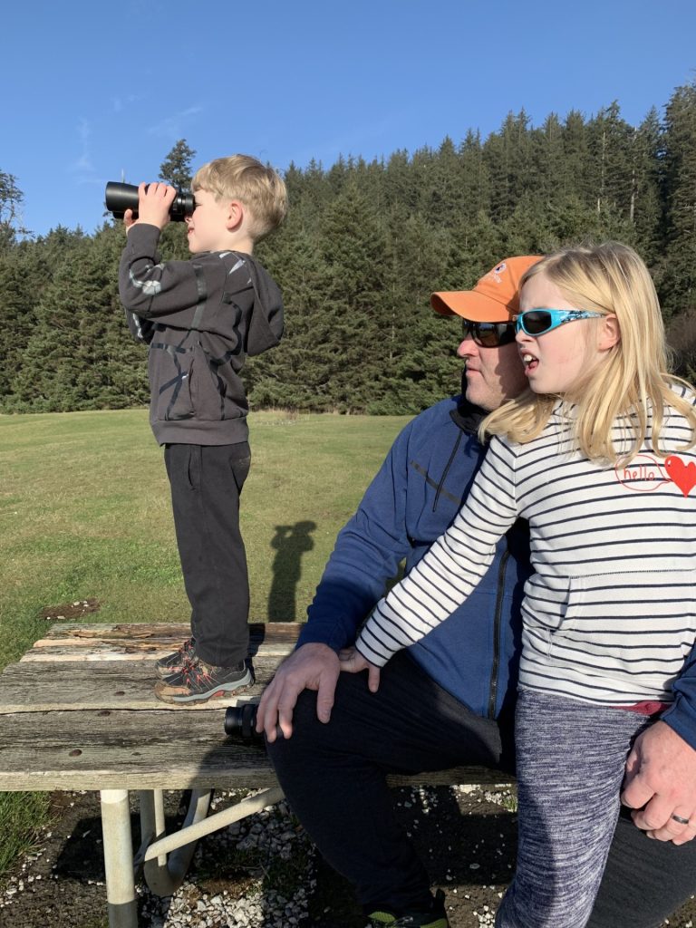 a dad sites on a park picnic table  with a young girl on his kneed. a young boy is standing on the table looking through binoculars