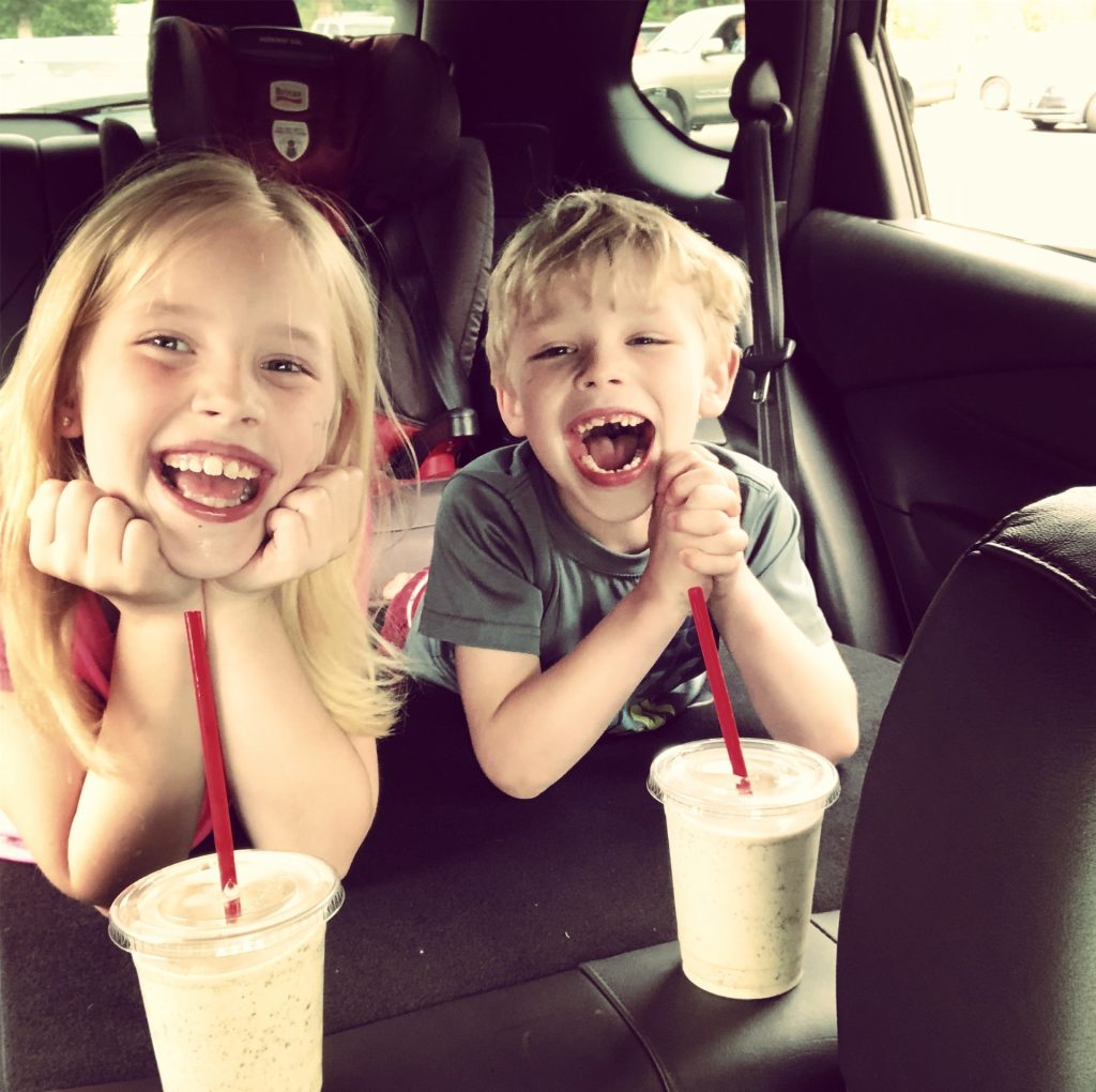 two kids in the back of a car smiling with drinks in front of them