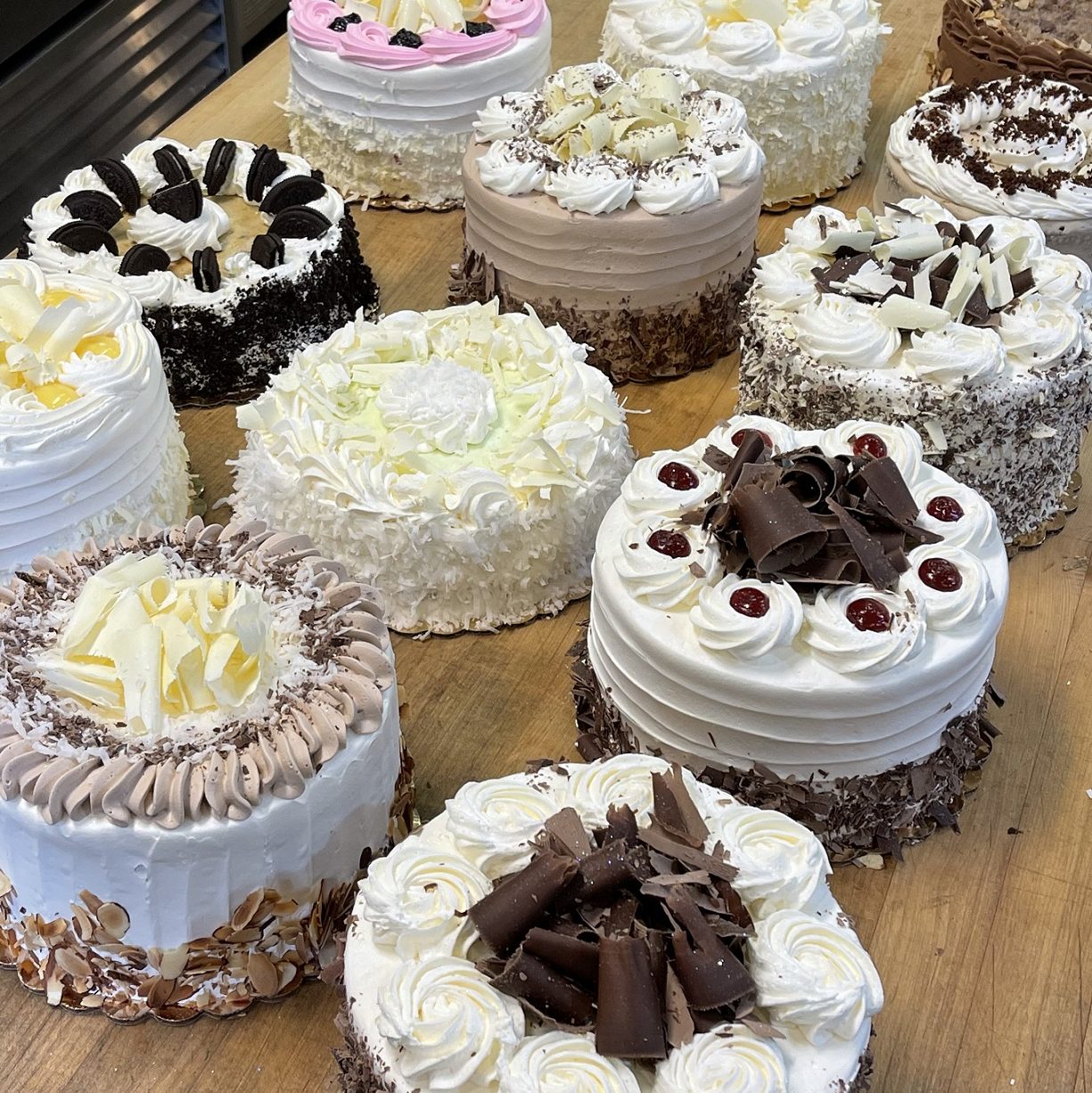 12 fancy cakes with all kinds of different decorations and frosting sit on a wooden kitchen counter. Some have cherries and chocolate shavings, others have white chocolate shavings, lots of frosting roses, and even slices of Oreos and coconut flakes
