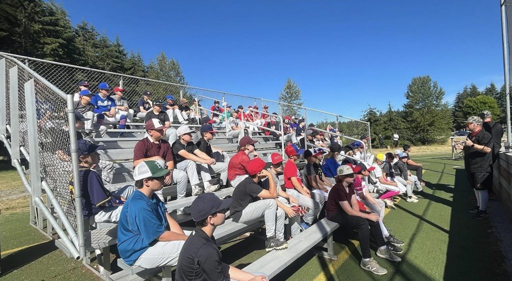 a bunch of kids in baseball hats and pants sit on bleachers outside . Two coaches in black stand facing them at Saint Martin's University