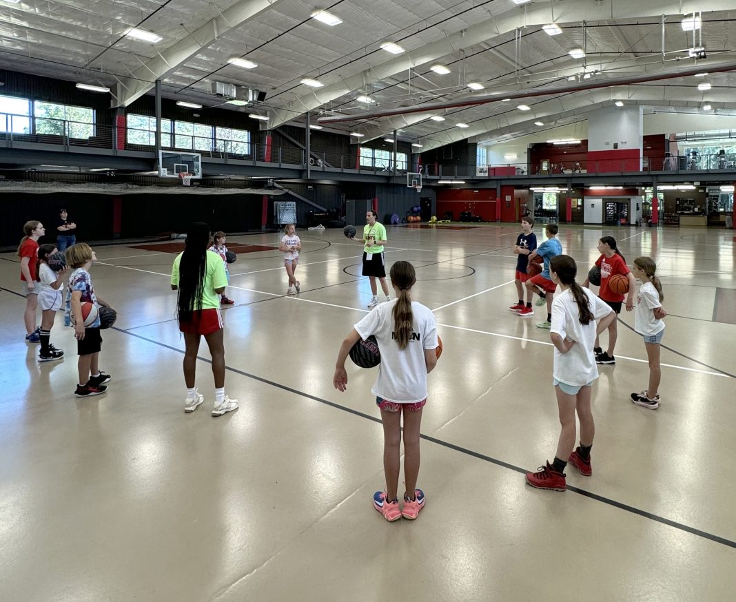 a group of kids in a circle on a basketball court at Saint Martin's University, some holding balls. A coach is talking to them, also holding a ball