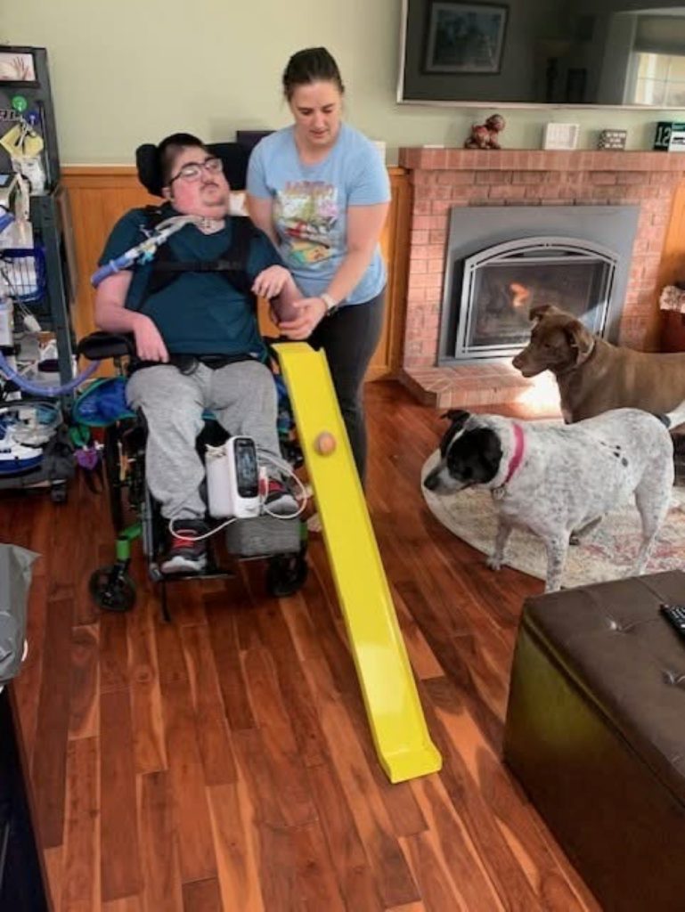 a man in a wheelchair drops a ball down a yellow slide attached to his chair with the help of a woman standing next to him. Two dogs are watching the ball
