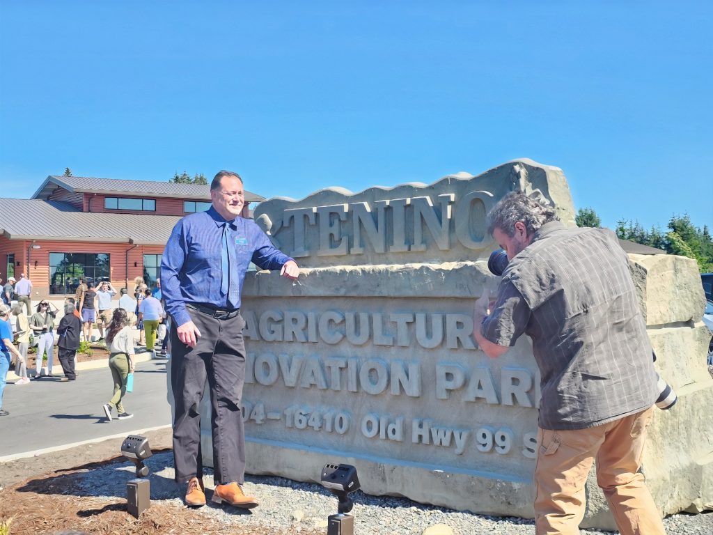 a man stands by a hand-carved stone sign, partially covering the words that say Tenino Agriculture Innovation Park 16410 Old Hwy 99 S. Another man takes a photo of him with a camera