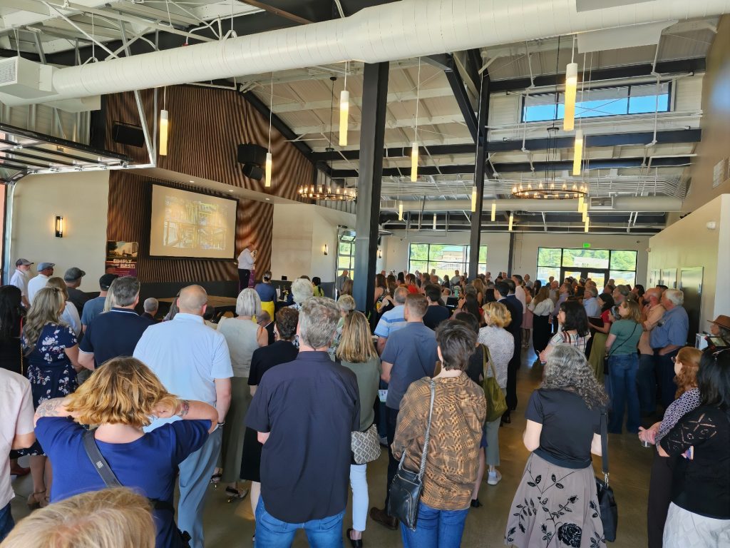 a large crowd stands inside the Tenino Agricultural Innovation Park for the grand opening, listening to a man on a stage with a large flat screen behind him
