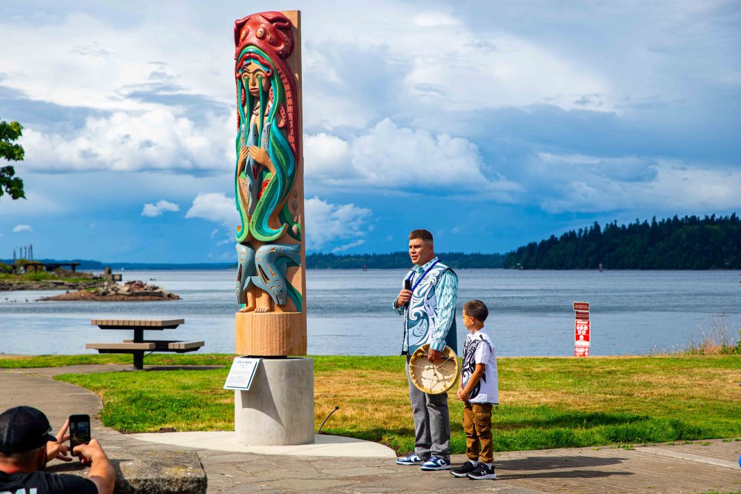 a man speaking into a microphone with a young boy next to him at West Bay Park, the new totem carving is in the background
