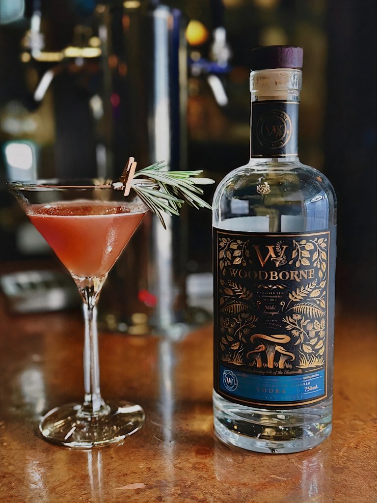 Woodborne Spirits bottle next to a martini glass with a pink liquid in it and a herb attached to rim with a tiny clothespin. both are sitting on bar top