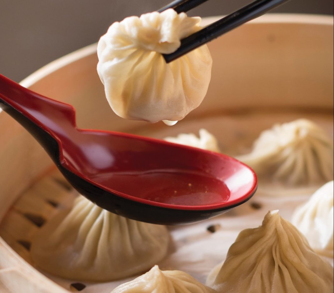 a bowl of bao with a red spoon with broth held over it. A bao is being held above the spoon with chopsticks