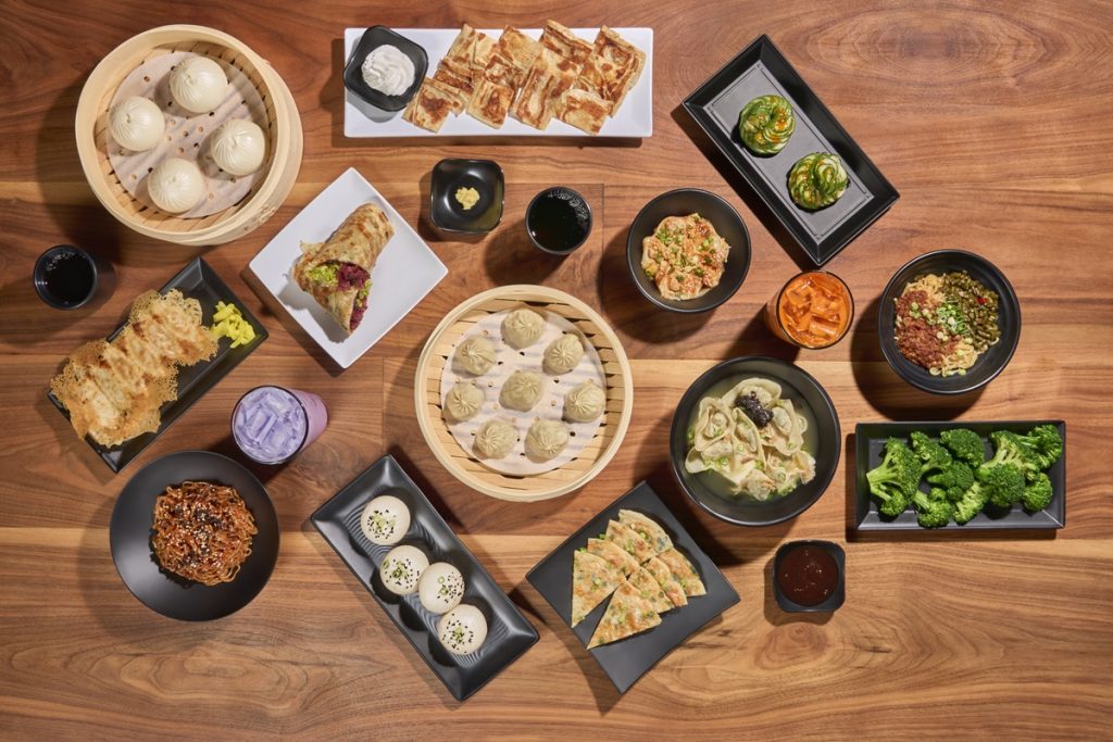 an aerial short of a bunch of dumpling, bao and noodle dishes with a purple drink in a glass, all sitting on a wood table at Dough Zone
