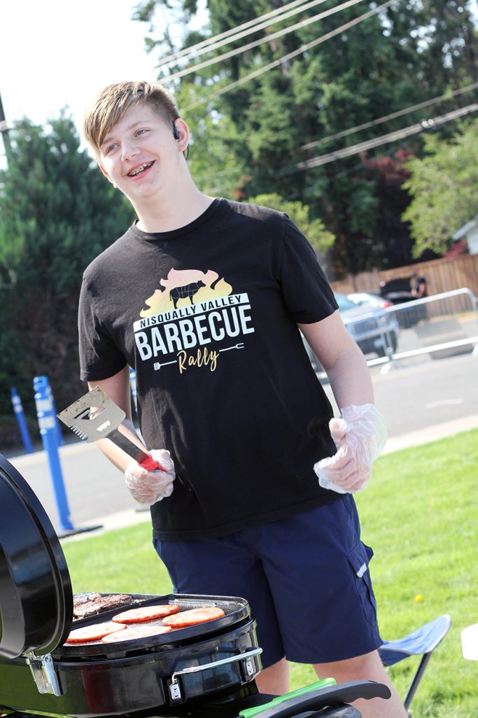 a young kid holding tongs smiles next to a grill with hamburger patties on it