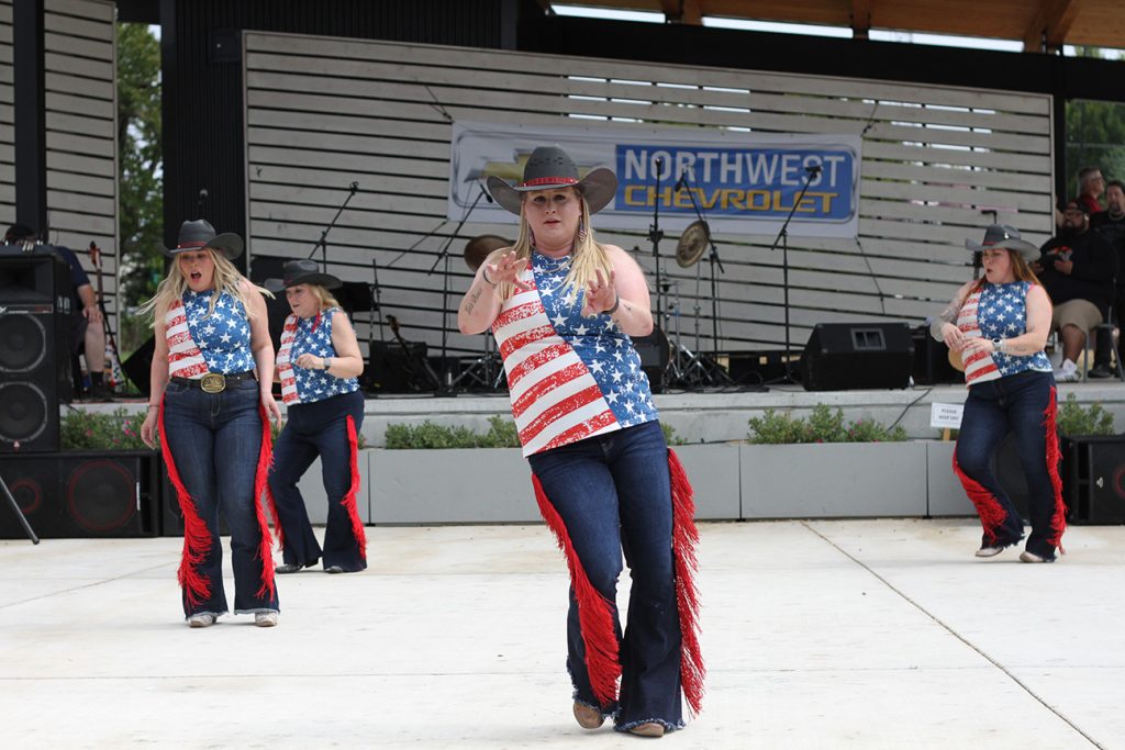 women with cowboy hats in red, white and blue American flag firsts and red and black pants dance and sing on a stage