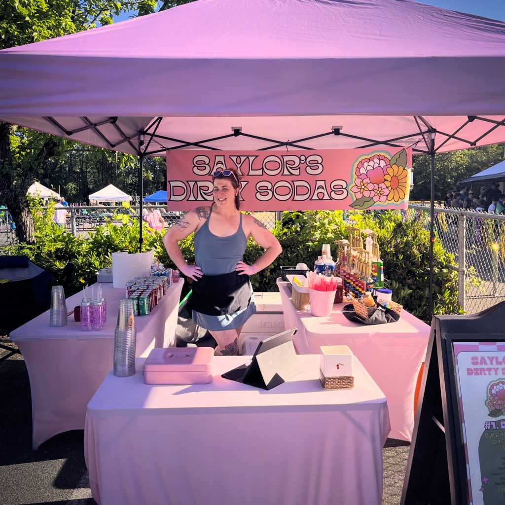 a woman with her hands on her bips stands behind tables in an U formation under a pink top up tent with a banner behind her that says Saylor's dirty Sodas