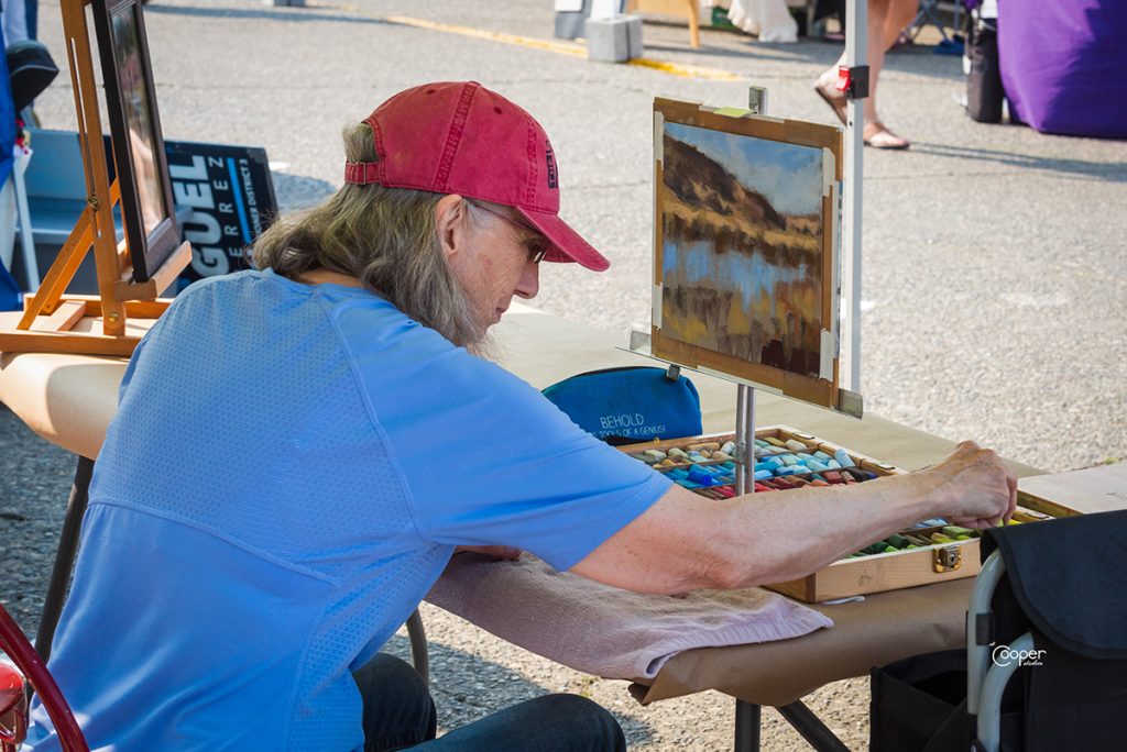 a woman in a t-shirt and a baseball hat sits at an easel drawing outside at an arts walk
