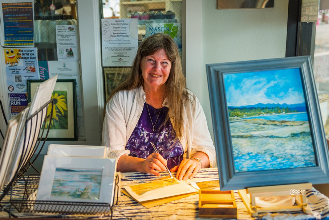 an artist sits at a table with her paintings on display