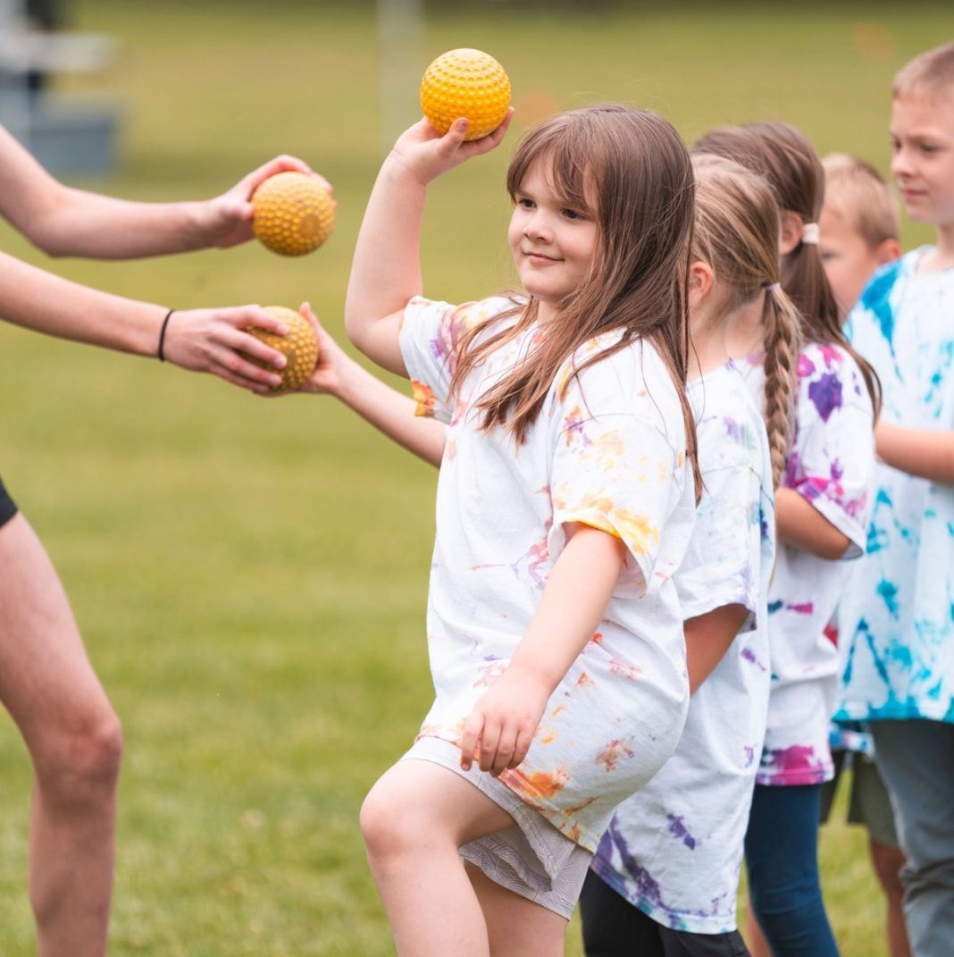young boys and girls in t-shirts and shorts stand in a line on a field, the hands of an adult are seen handing the first two kids orange balls. The kid at the front of the line is throwing her ball.,