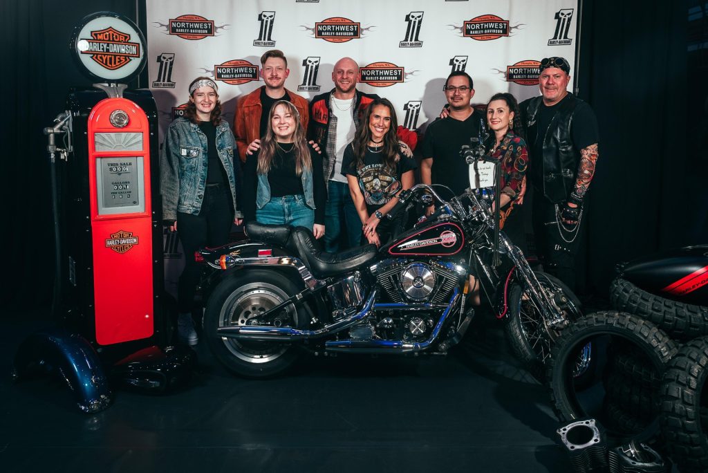 4 men, two women and two teenagers smile for a photo behind a Harley Davidson motorcycle. A Harley Davidson fuel pump is next to them, with tires pile don the other side and a step-and-repeat with the Northwest Harley Davidson logo on it is behind them.