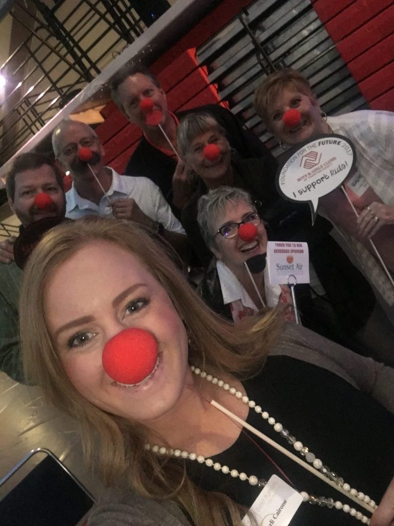 Adults wearing red clown noses and holding up fake mustaches and glasses on wooden dowels smile for the camera
