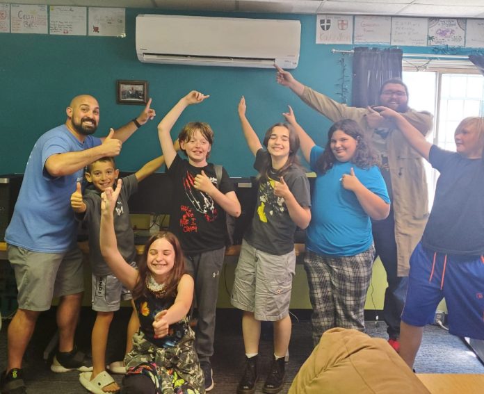 a group of kids and two adults stand under a ductless A/C system, pointing to it with one hand and giving a thumbs up with the other.