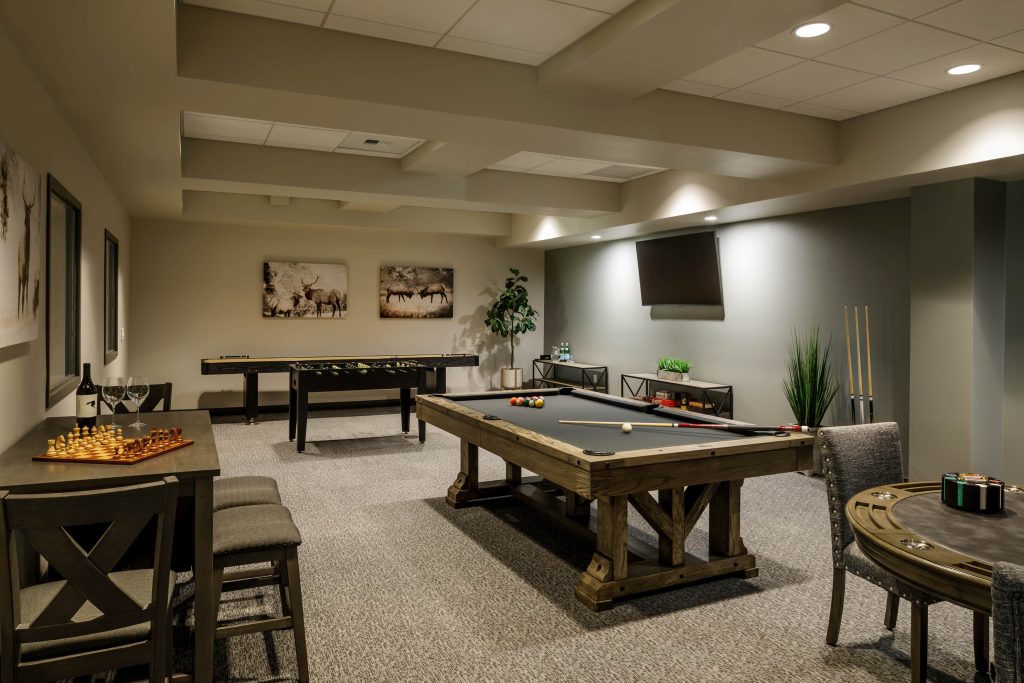 a room with a pool table, foosball, a table with a chess board and a bottle of wine with two glasses, and a poker table with a poker chip holder on it