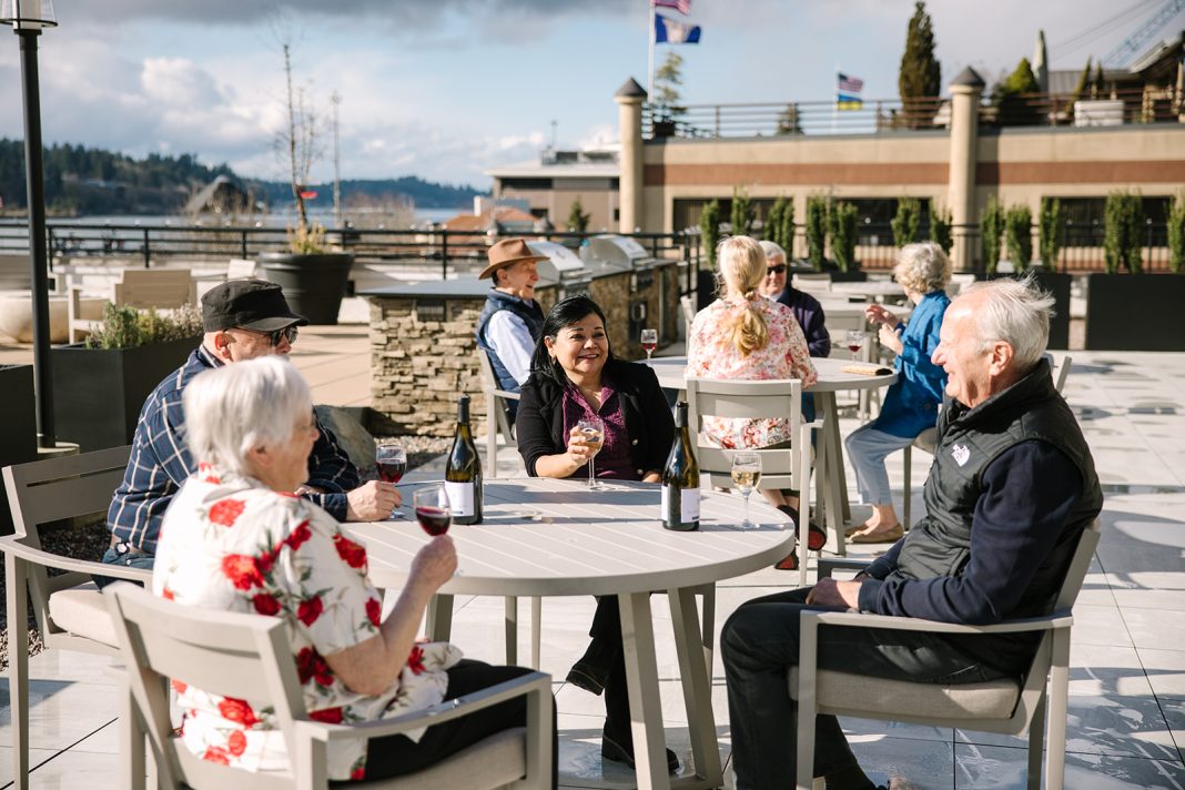 a group of senior citizen sitting at round tables on a terrace. There are wine bottles and glasses of wine on the tables
