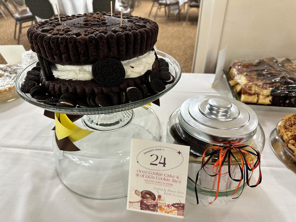 a chocolate cake made to look like a giant Oreo sits on a glass cake platter on a table with an empty glass cookie jar. A card next to it with the number 24 and the words "Oreo cookie cake & 1 of GG"s cookie jars"