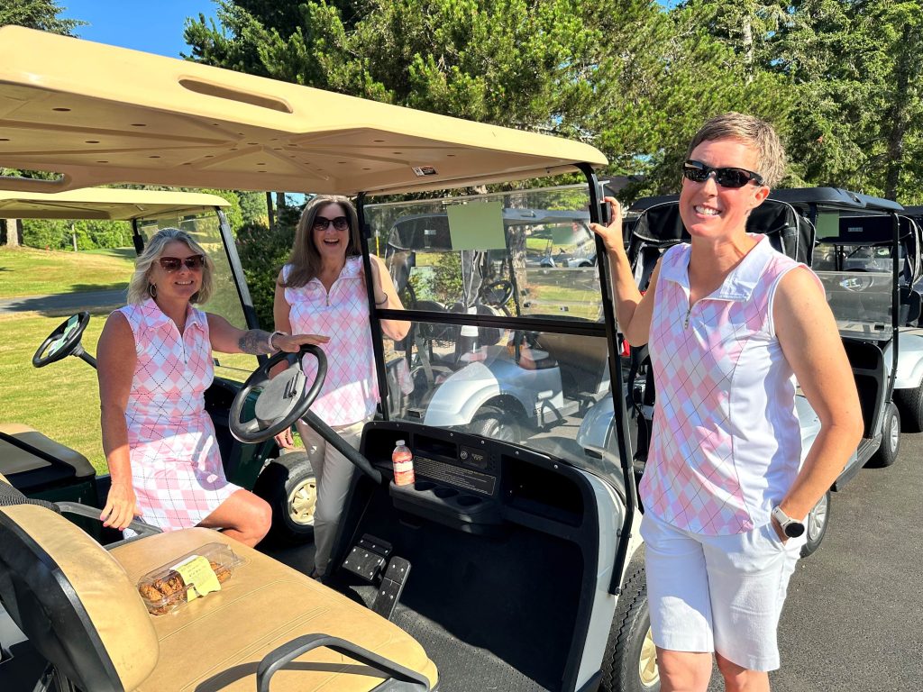 There women in matching white and pink argyle sleeves collared shirts smile next to a golf cart. There are other golf carts around them.