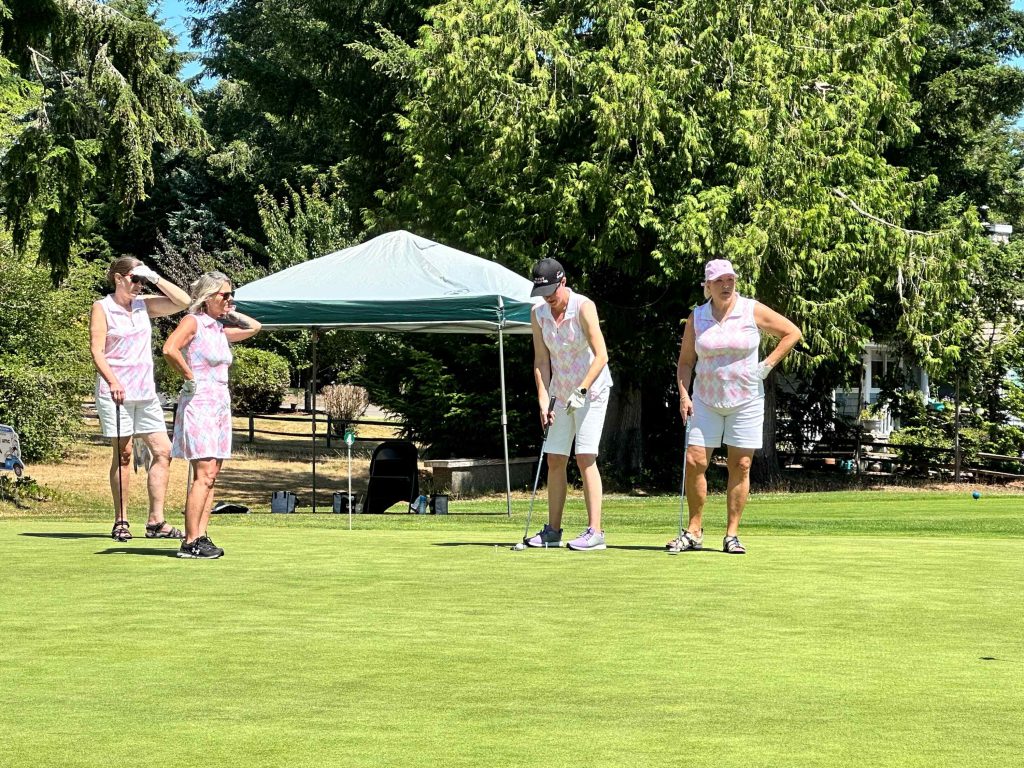 Four woman on a golf course, one is about to swing her club. A pop-up tent is behind them