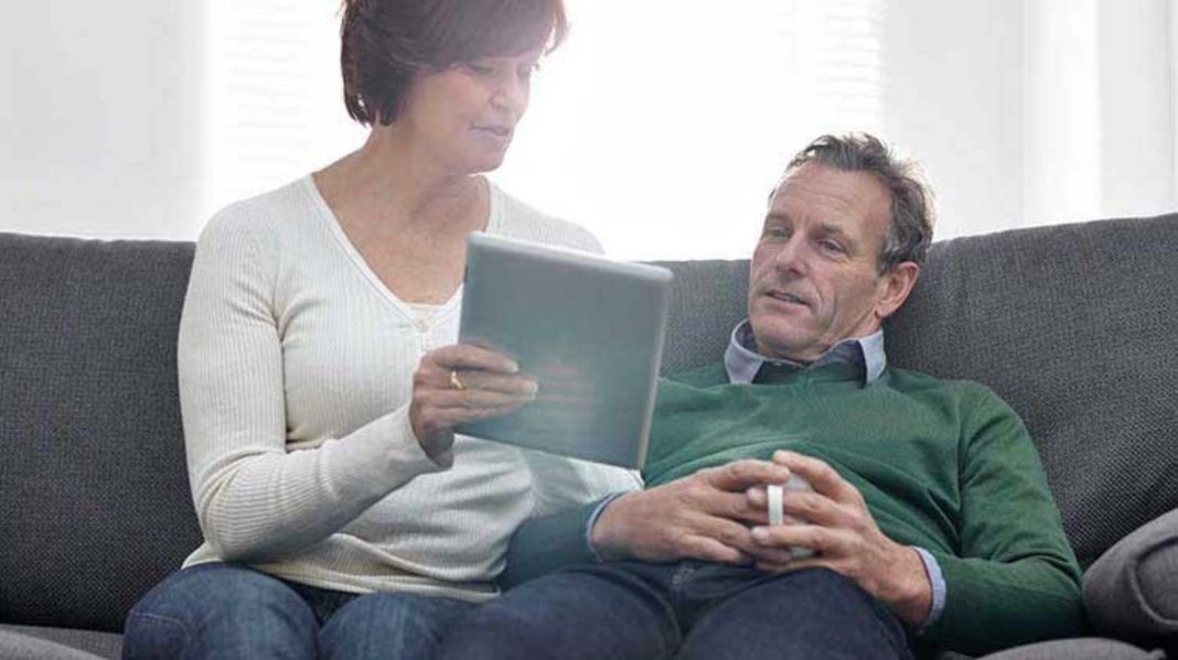 a man and woman sitting on a couch. The woman is a tablet up and they are both looking at it. The man is holding a cup of coffee.