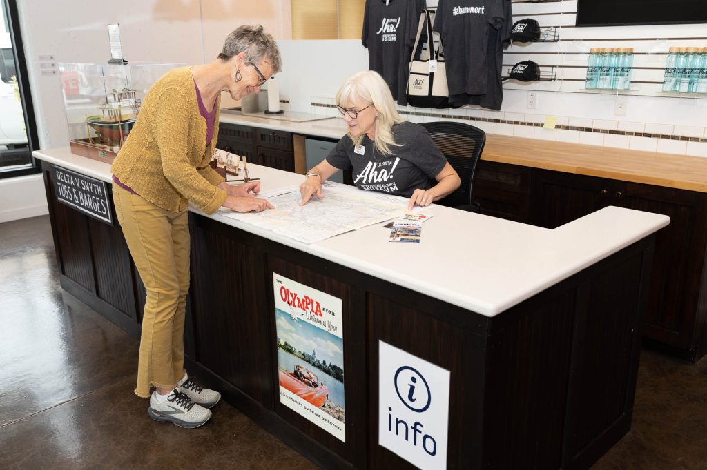 a woman leans over a counter at the Olympia visitor center and looks at a map with the woman sitting behind the counter, both of them are pointing to spots on the map with an index finger