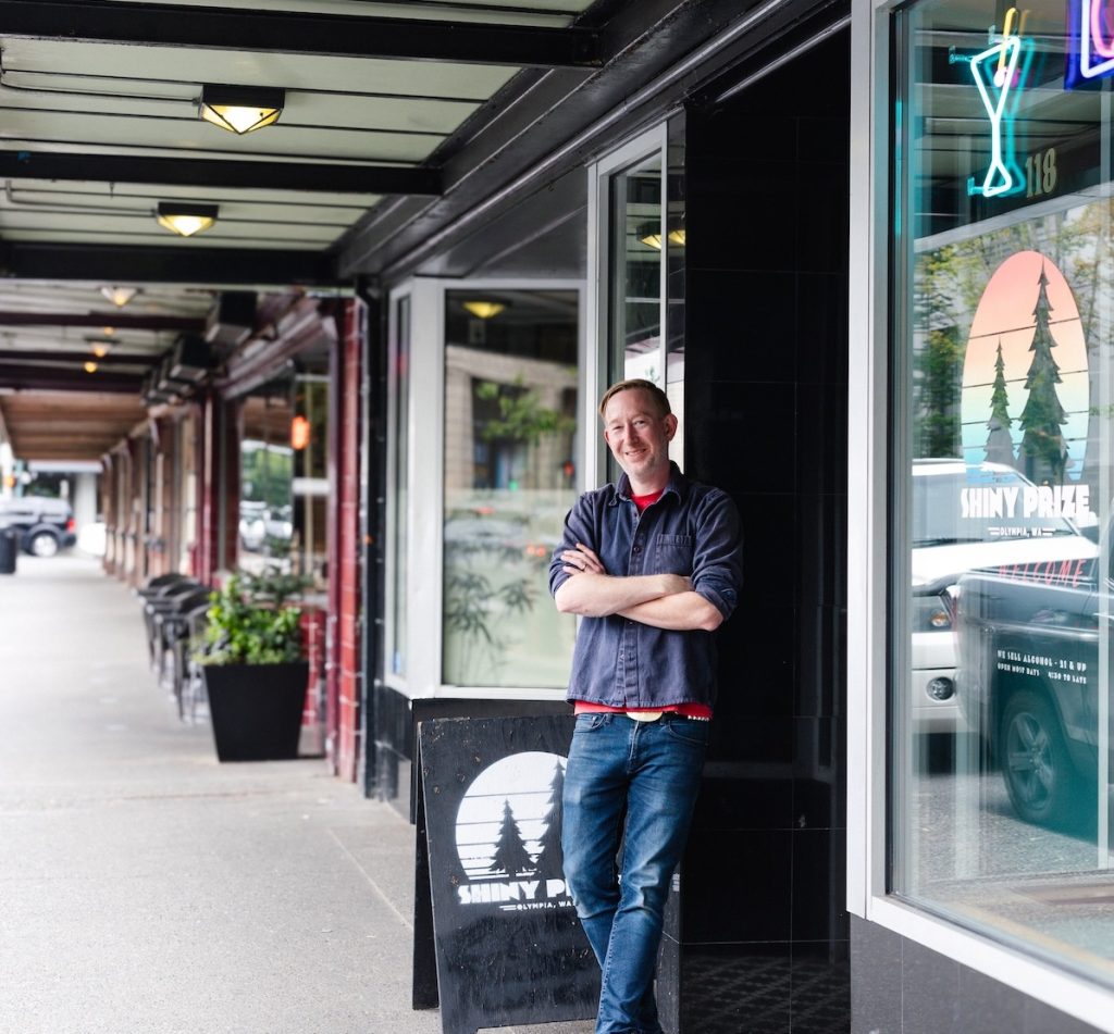 Shiny Prize owner Tim Zohn leans against the doorway of the bar in a button up shirt and jeans