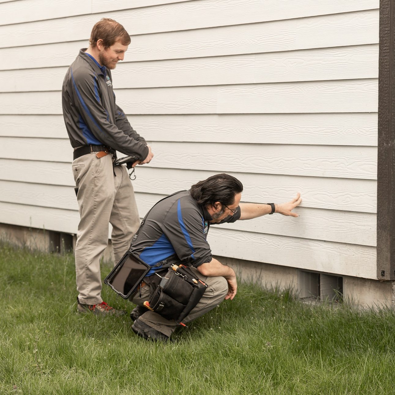 Two Boggs home inspection technicians examine the siding and crawl space of a house, one is standing and the other is crouching with his hand on the siding