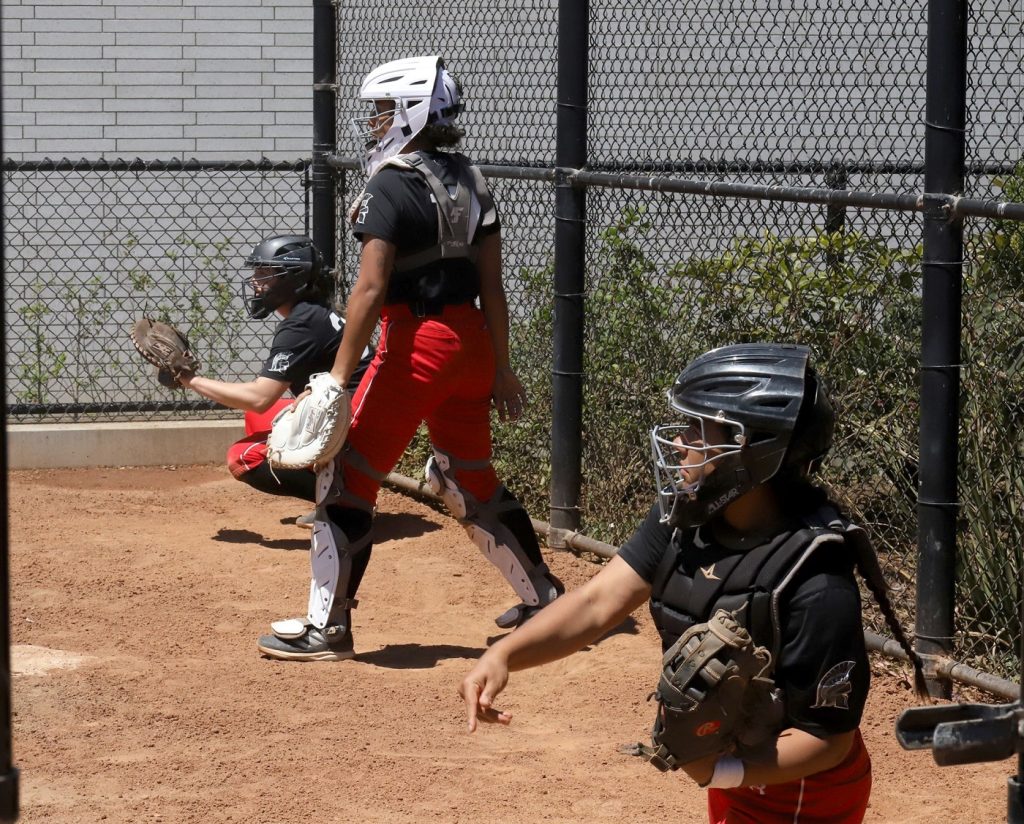 e Saint Martins Softball players in black and red uniforms and catcher helmets throwing balls in a dirt area