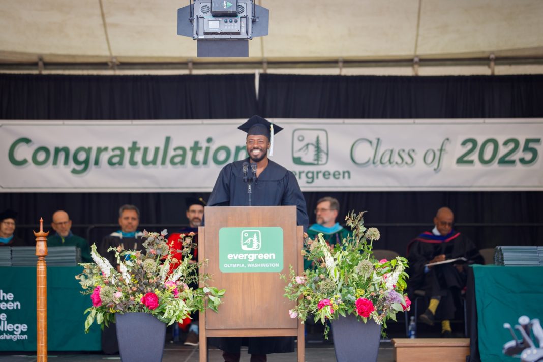 Humanitarian and influencer Kwabi Amoah-Forson stands at a podium with staff from Evergreen State College sitting behind him. a Banner says "Congratulations Class of 2025 - Evergreen" on the wall behind him