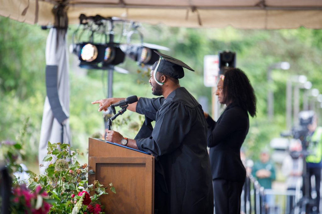 Humanitarian and influencer Kwabi Amoah-Forson stands at a podium with woman standing next to and slightly behind him. Photo is taken from the side of the stage