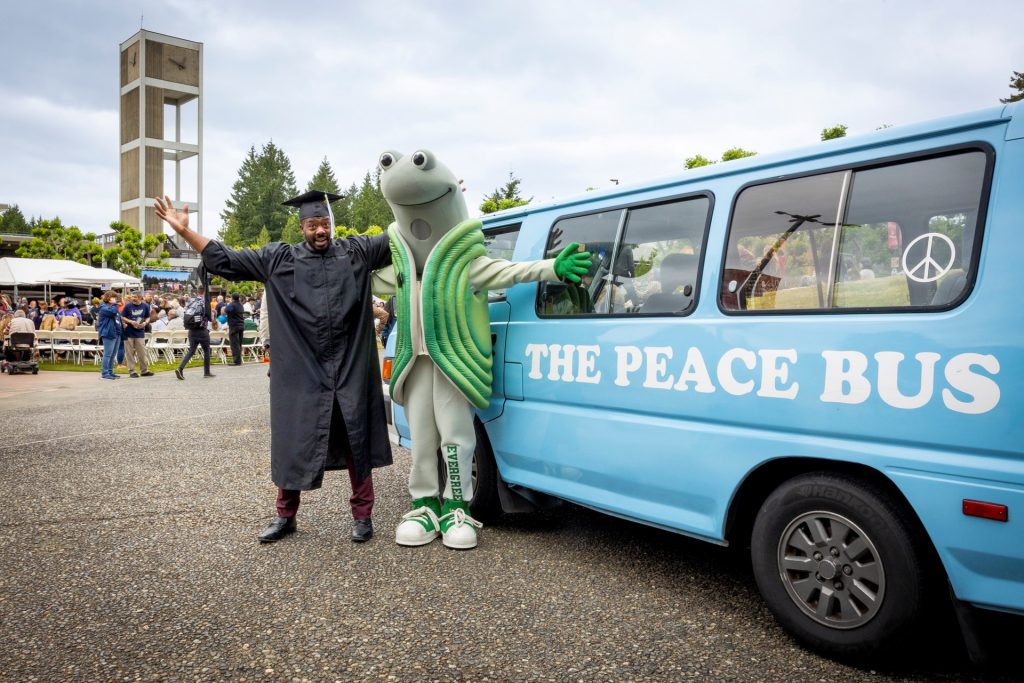 Kwabi Amoah-Forson in a graduation cap and gown stands in front of The Evergreen State College's mascot, Speedy the Geoduck, in front of The Peace Bus. Photo courtesy: The Evergreen State College
