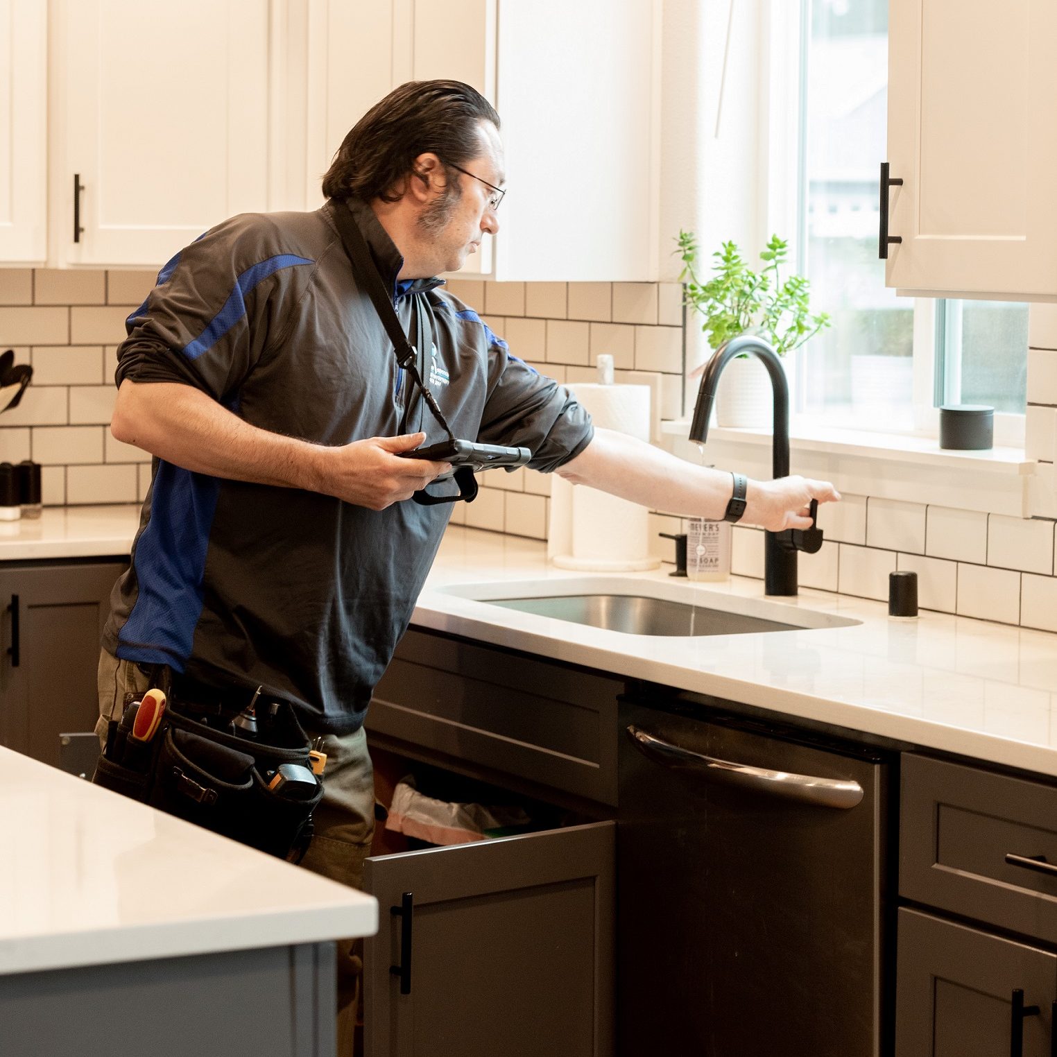 A Boggs home inspection technician tests a water faucet in a kitchen, the under sink cabinet door is open