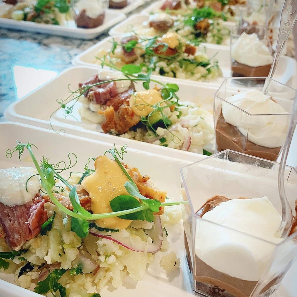 small white dishes with some type of rice, meat, sauce and veggie dish next to a cup with chocolate mousse topped with whipped cream and a spoon inside it.