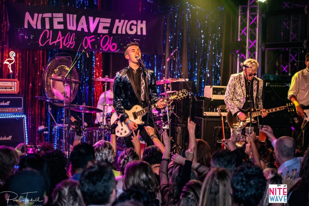 A rock band, Nite Wave, on a stage playing to a crowd of people. A banner with the band's name is behind them.
