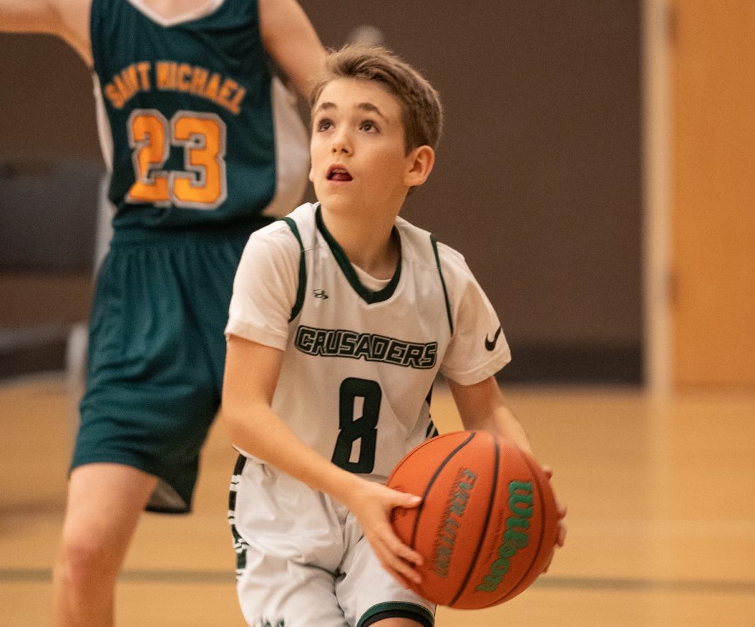little boy running on a basketball court with a basketball in an Evergreen Christian School jersey. Another boy in a different green uniform is behind him