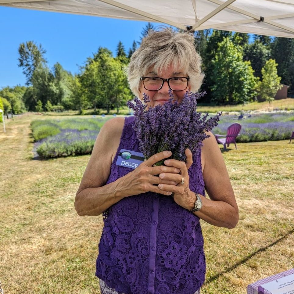 Peggy wearing a purple sleeveless shirt, holding up a bouquet of dried lavender with the Evergreen Valley Lavender Farm fields in the background.