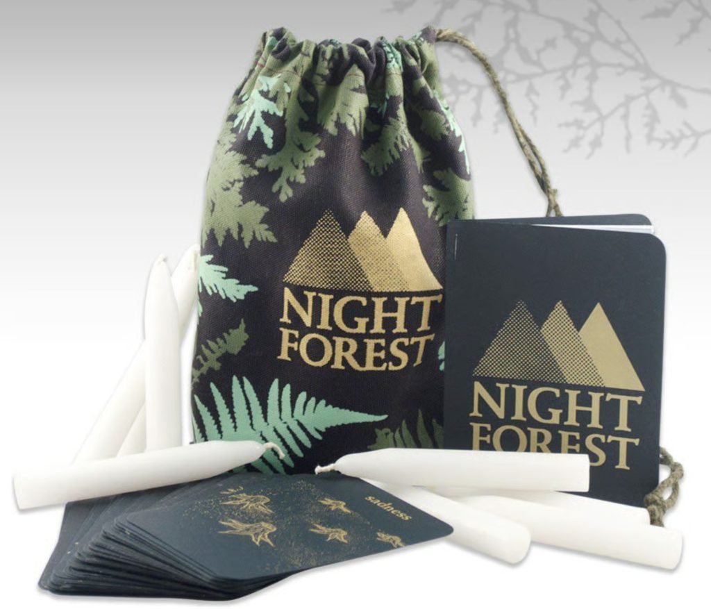 a drawstring bag and book with tree branches and the words "Night Forest" on them. white taper candles are on their side laying next to them, along with a green card deck, fanned out in front