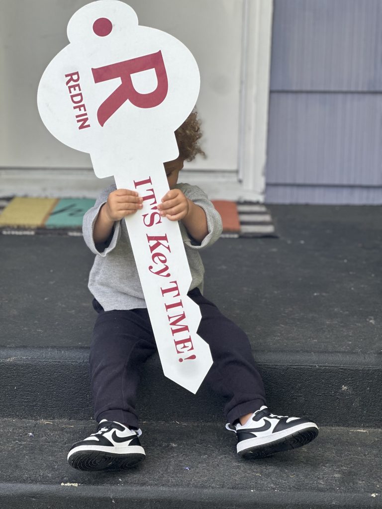 a little kid sits on a porch step of a house holding a giant key-shaped sign in front of them. The white key says "R Redifin: it's key time" on it in red letter