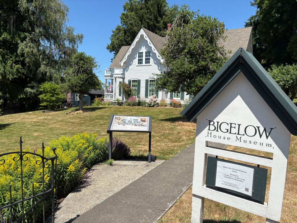 The Bigelow House Museum with a house shaped sign out front that says 'Bigelow House Museum' and an informational guide a bit further down the sidewalk that leads up to the white house with green shutters