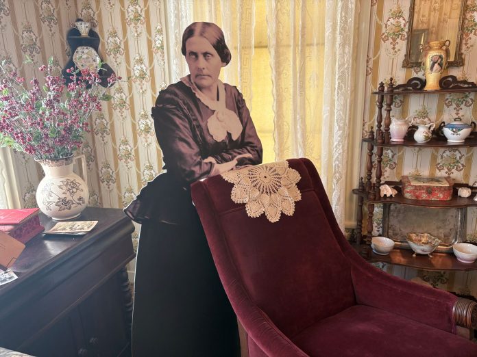 A life-size cardboard cutout of reformer Susan B. Anthony stands next to the red velvet chair she reportedly sat in at the Bigelow House. A desk with flowers and pamphlets are behind the cut out and a tied shelf is next to the chair with different porcelain vases and pitchers on it.