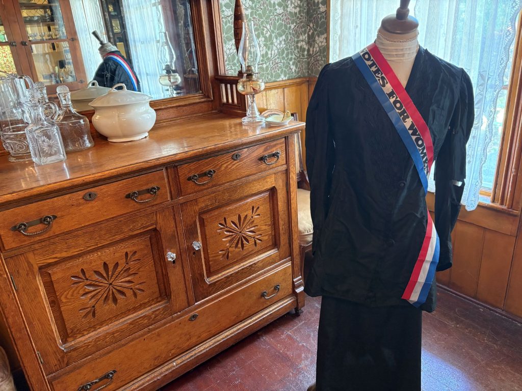 a Woman's black dress on a dress form with a red, white and blue striped sash with the words " Votes for Women" Lies across it diagonally from one shoulder. A chest with a mirror is behind it. Glass canisters and pitchers are on the chest top.