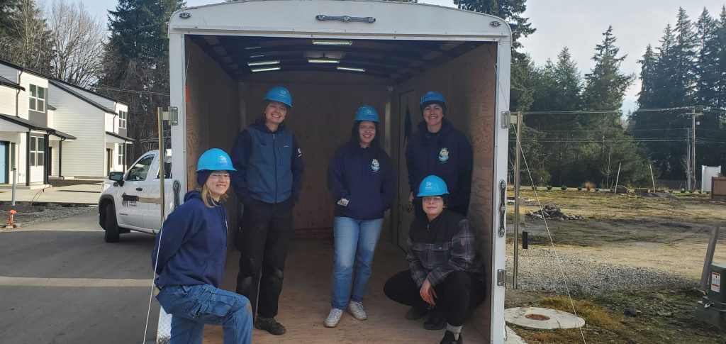 Five women in hardhats stand inside a covered trailer attached to a truck