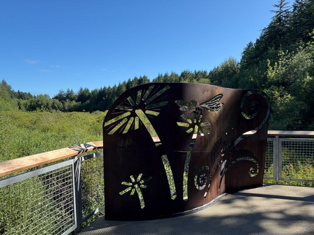 A large bronzed piece of metal with flower and dragon fly cut outs, stand up on a metal boardwalk railing at Grass Lake Nature Park in Olympia 