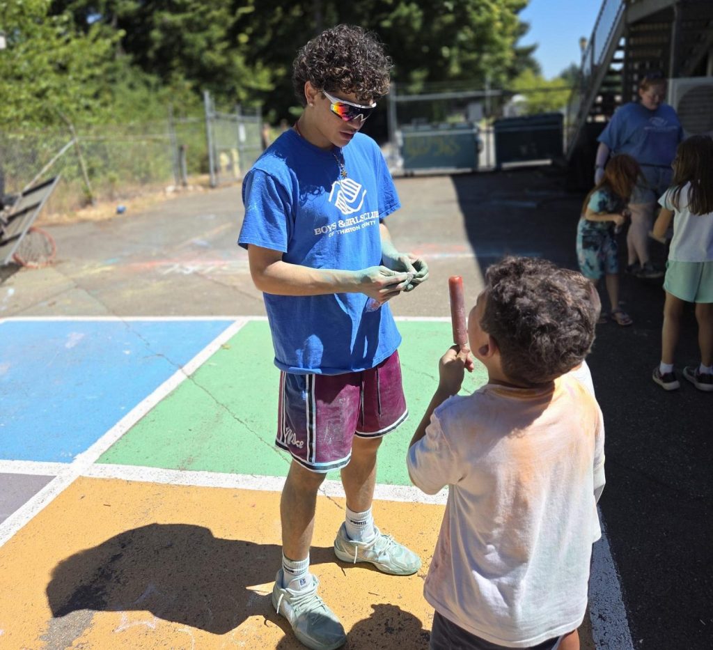 an young adult in a Boys & Girls Clubs shirt giving a popsicle to a child as they stand on a chalk covered concrete playground