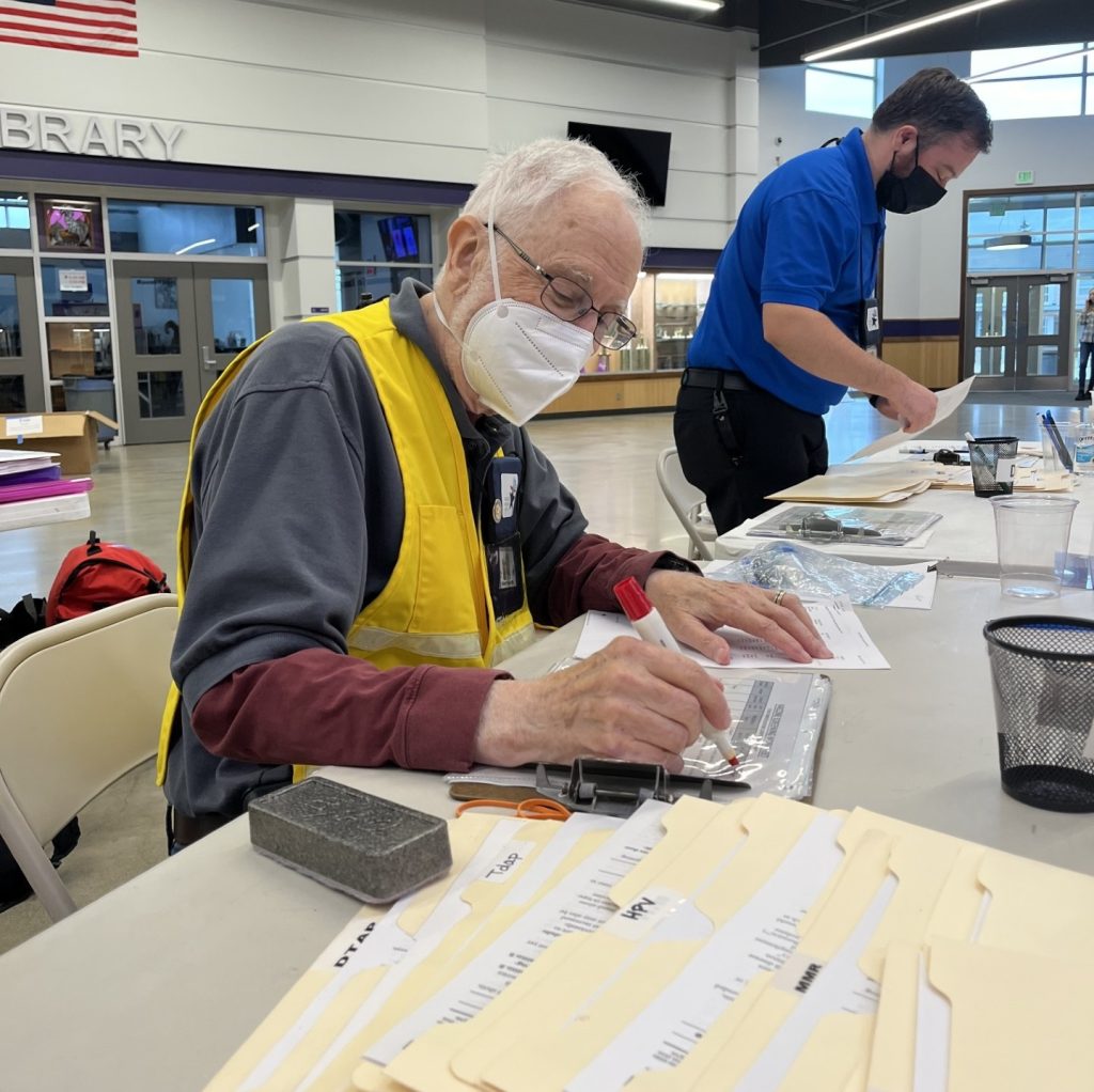 a man in a face mask sits at a table with a bunch of file folders and other paperwork. A man further down the table is standing up looking at papers on the table